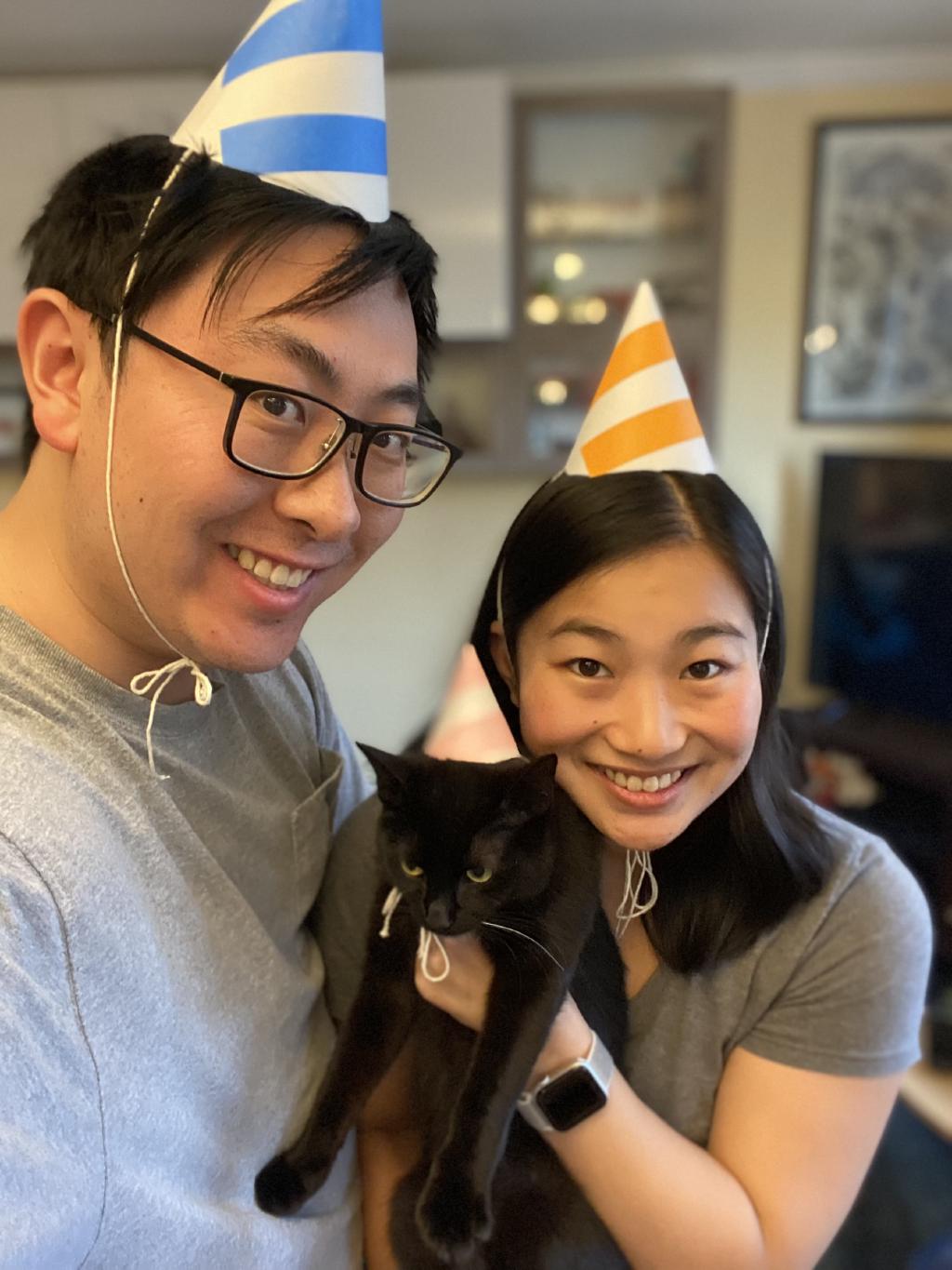 Kelly and Eric holding Moon Moon, one of their cats, and wearing handmade party hats